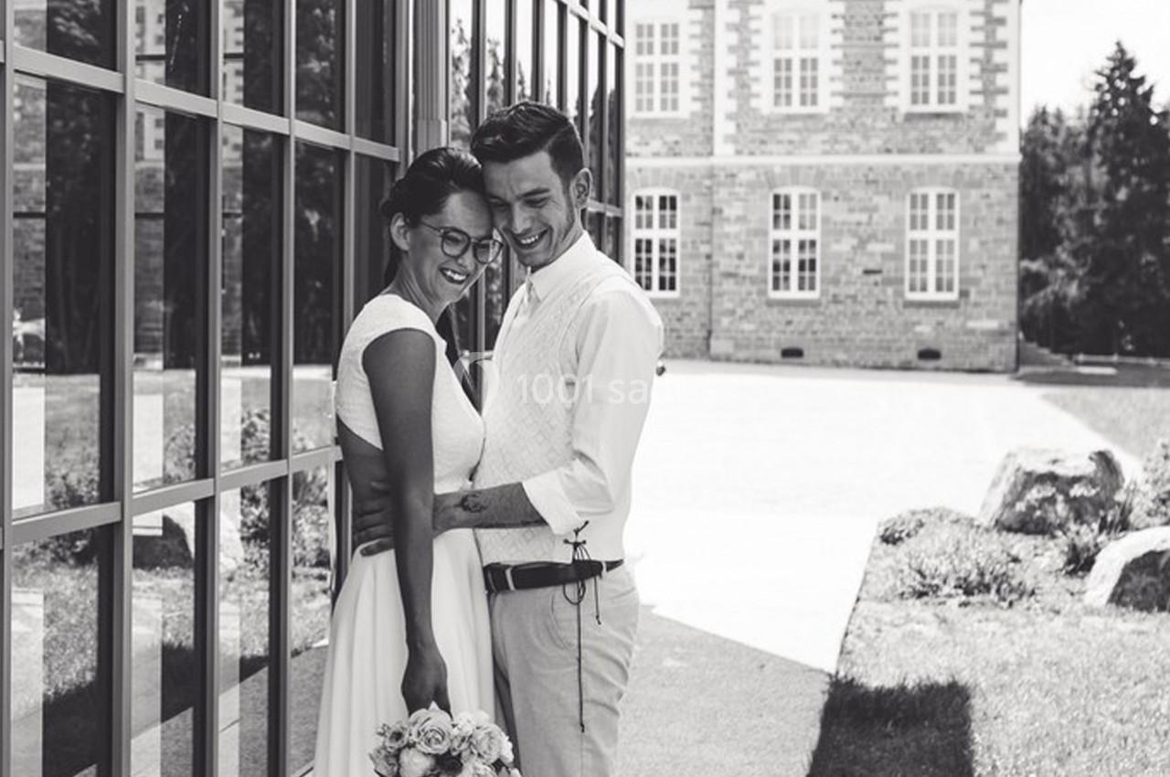 Un couple souriant en tenue de mariage pose devant un bâtiment vitré et un espace extérieur arboré.