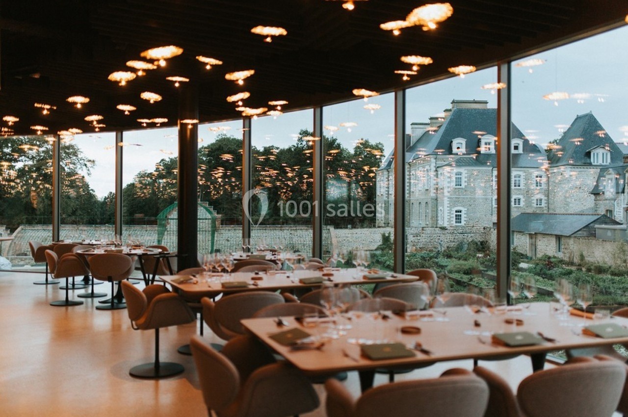 Salle de restaurant moderne avec grandes baies vitrées donnant sur un jardin et un bâtiment en pierre.