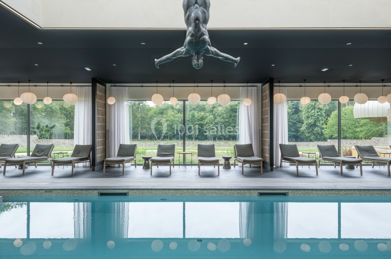 Piscine intérieure avec chaises longues alignées, grandes baies vitrées donnant sur un jardin et sculpture suspendue.