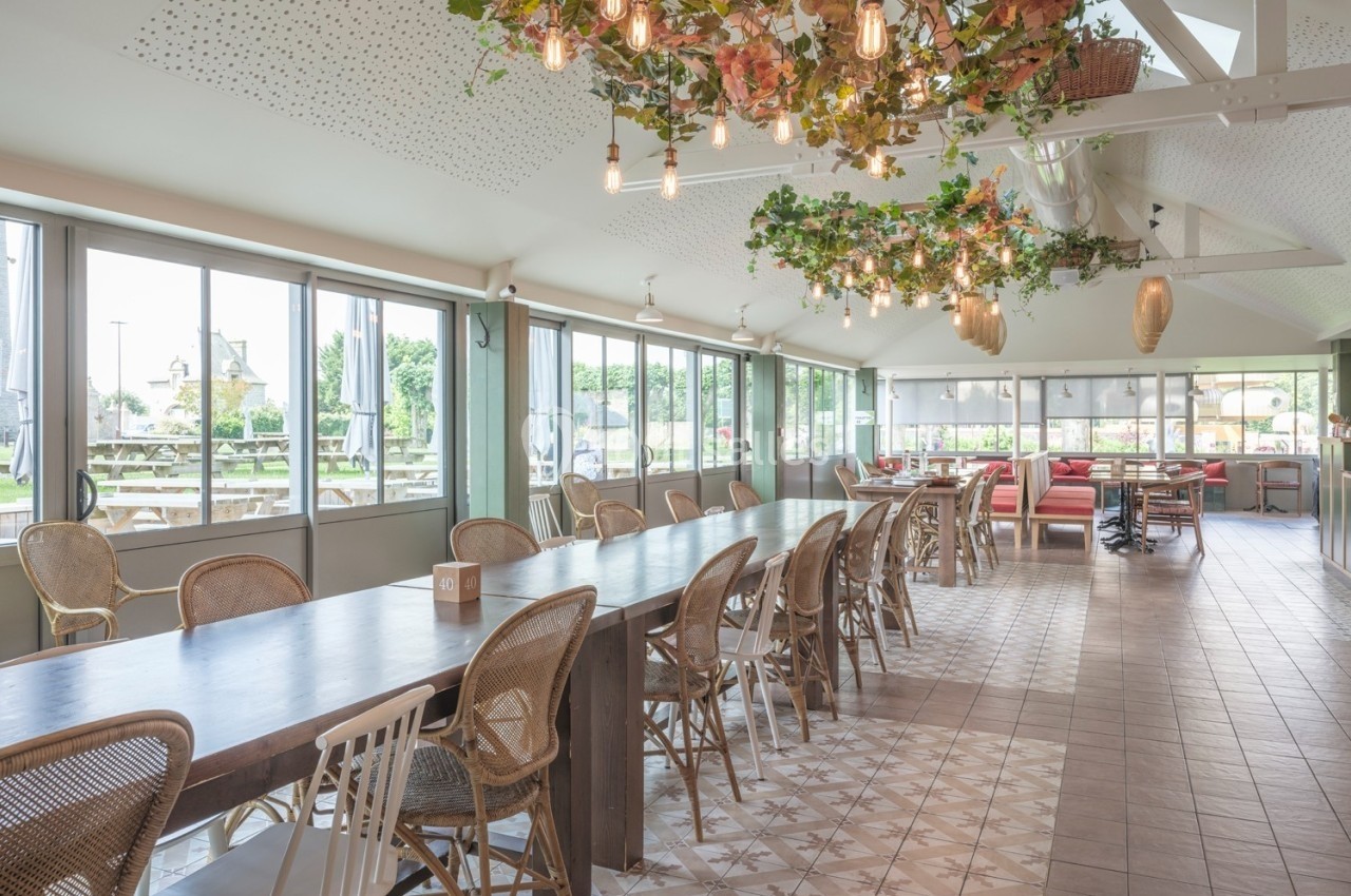 Salle lumineuse avec de grandes baies vitrées, tables en bois, chaises en osier et décoration florale suspendue.