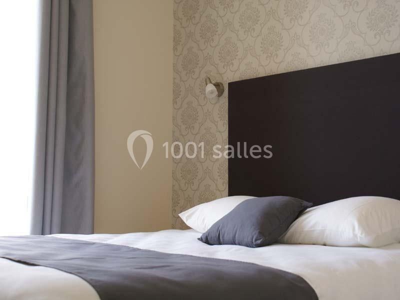 Lit double avec tête de lit sombre, draps blancs, coussins gris et mur tapissé à motifs dans une chambre lumineuse.