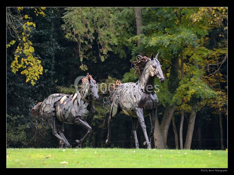 Deux sculptures de chevaux en métal courant sur une pelouse, avec des arbres en arrière-plan.