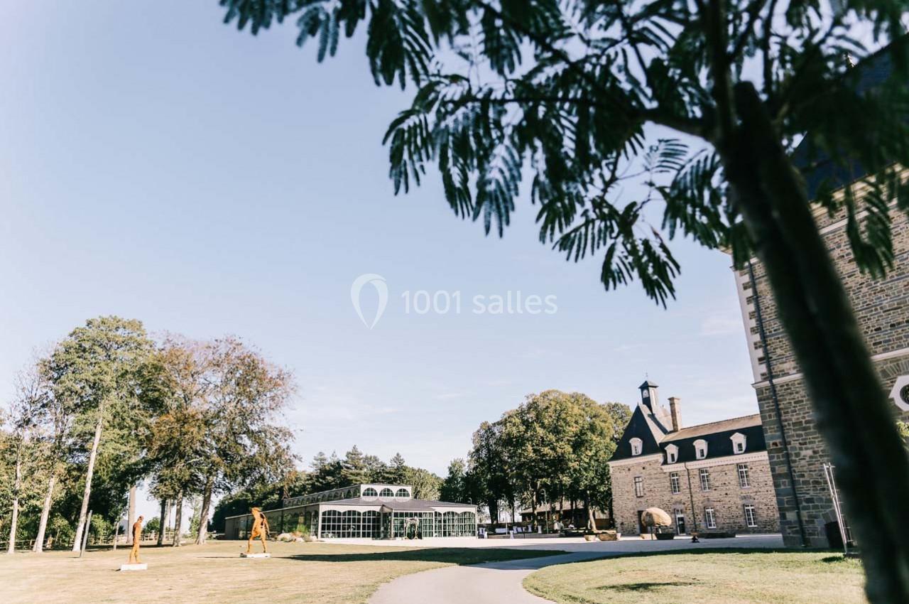 Vue d'un bâtiment en pierre avec une serre vitrée, entouré d'arbres et d'une pelouse sous un ciel dégagé.
