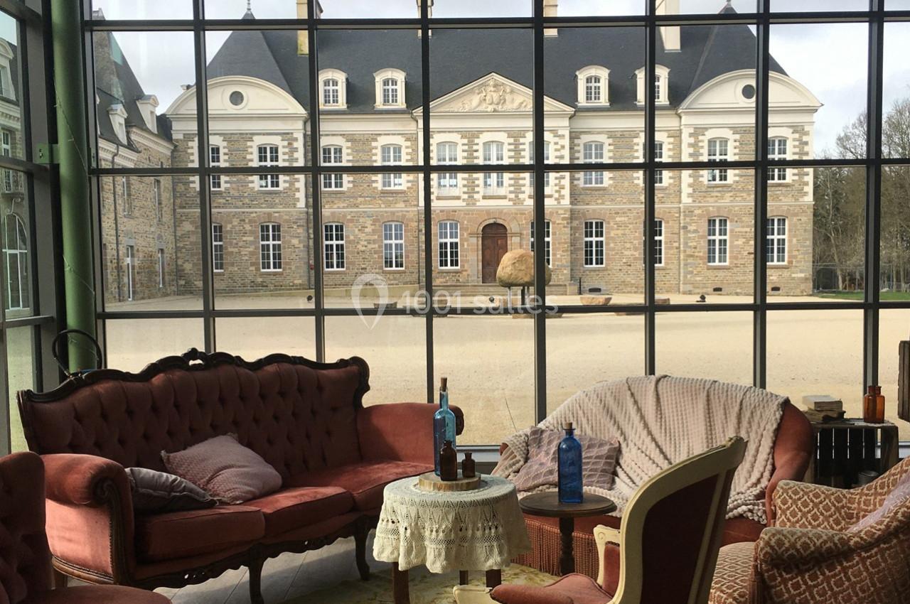 Salon avec mobilier ancien et vue sur une façade de château à travers de grandes baies vitrées.