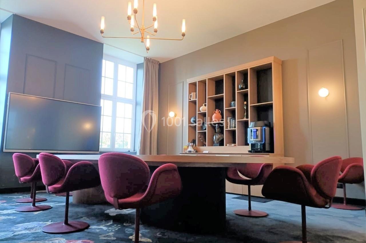 Salle de réunion lumineuse avec table en bois, chaises rouges, étagères décoratives et grande fenêtre.