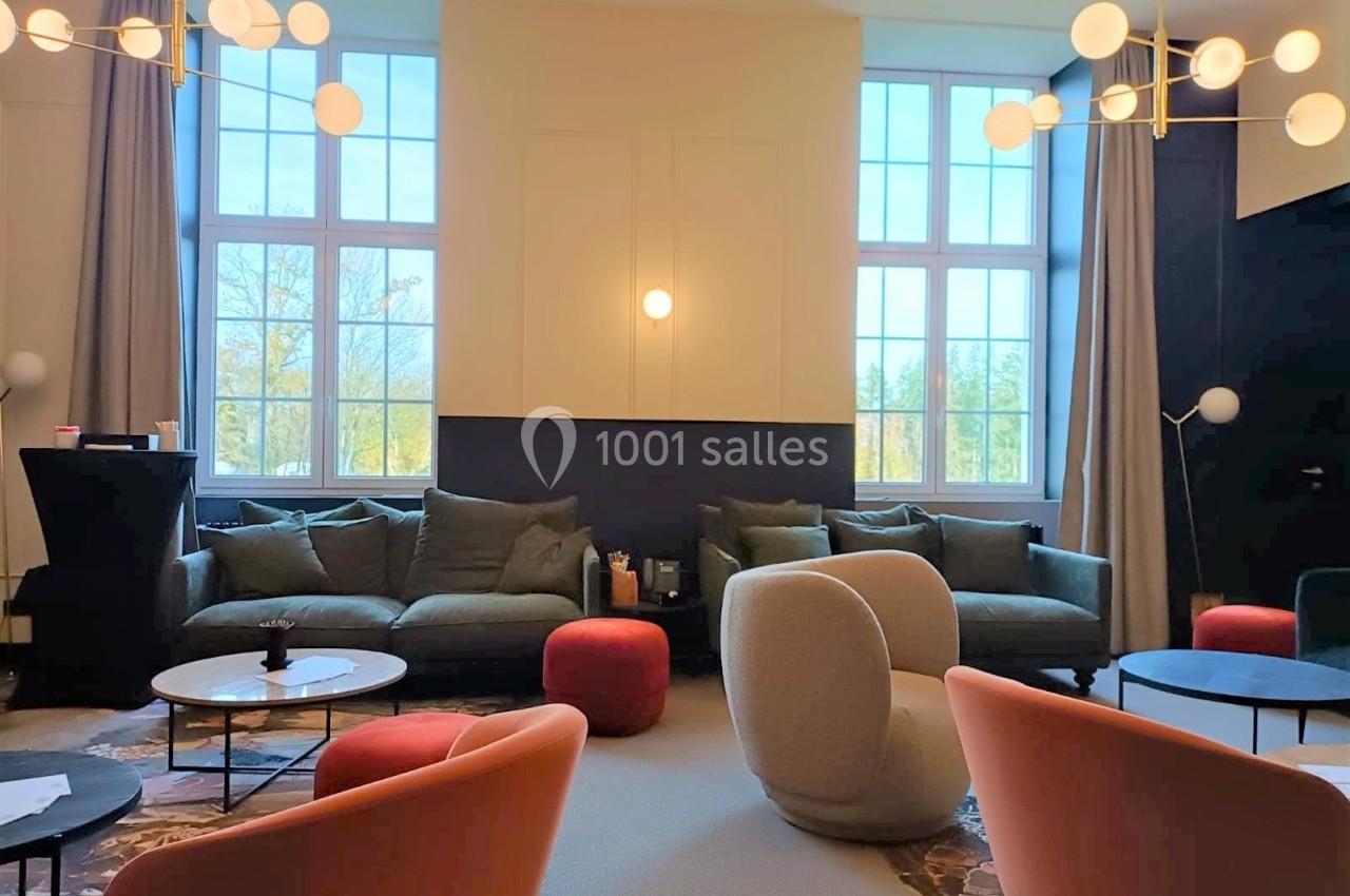 Salon lumineux avec grandes fenêtres, canapés verts, fauteuils modernes, tables basses et luminaires suspendus.