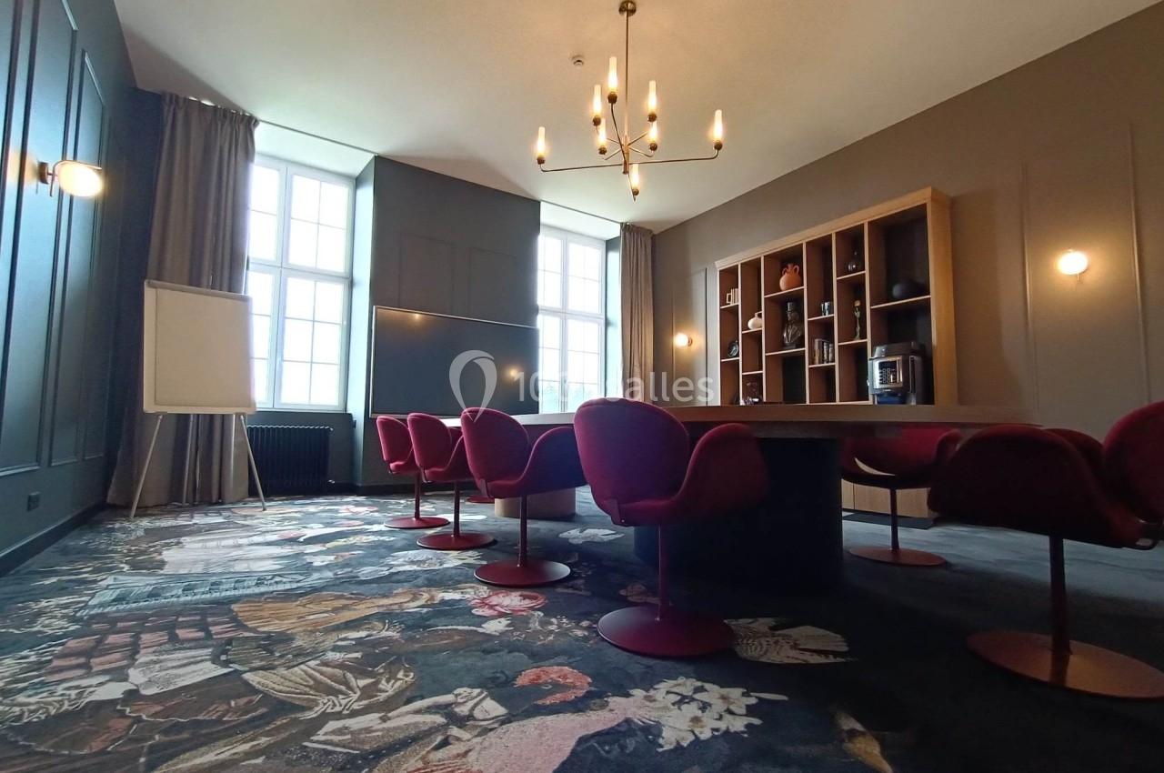 Salle de réunion avec table ovale, chaises rouges, tableau blanc, étagères en bois et lustre suspendu.