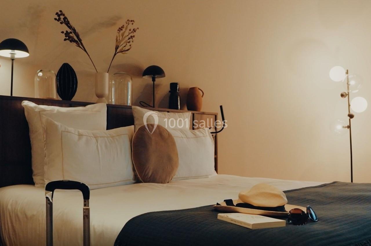 Lit double avec coussins et couverture, valise au pied, chapeau et livre posés, éclairage doux et décor minimaliste.