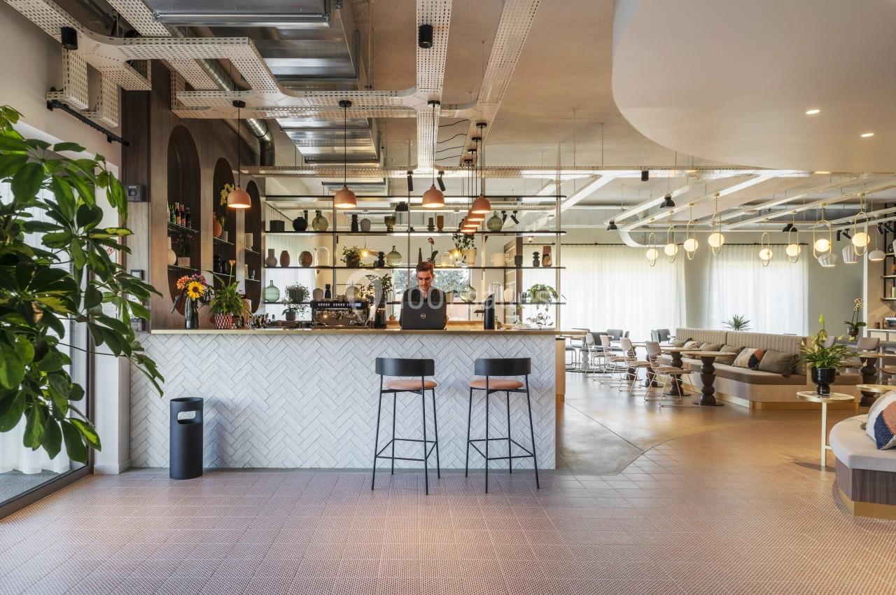 Espace lumineux avec un comptoir de bar, des tabourets, des plantes et un homme travaillant sur un ordinateur portable.