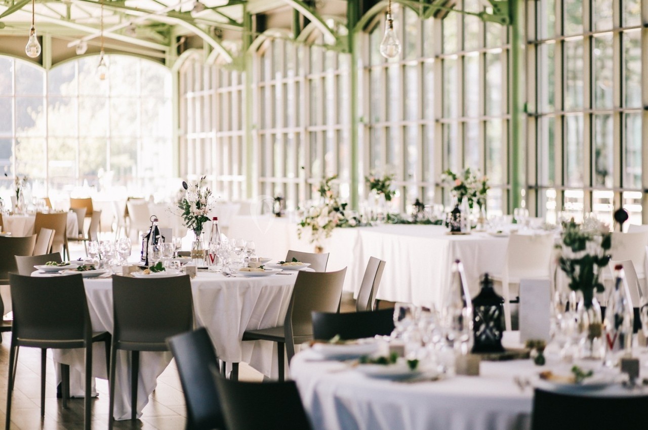Salle lumineuse avec grandes baies vitrées, tables rondes dressées avec nappes blanches et décorations florales.