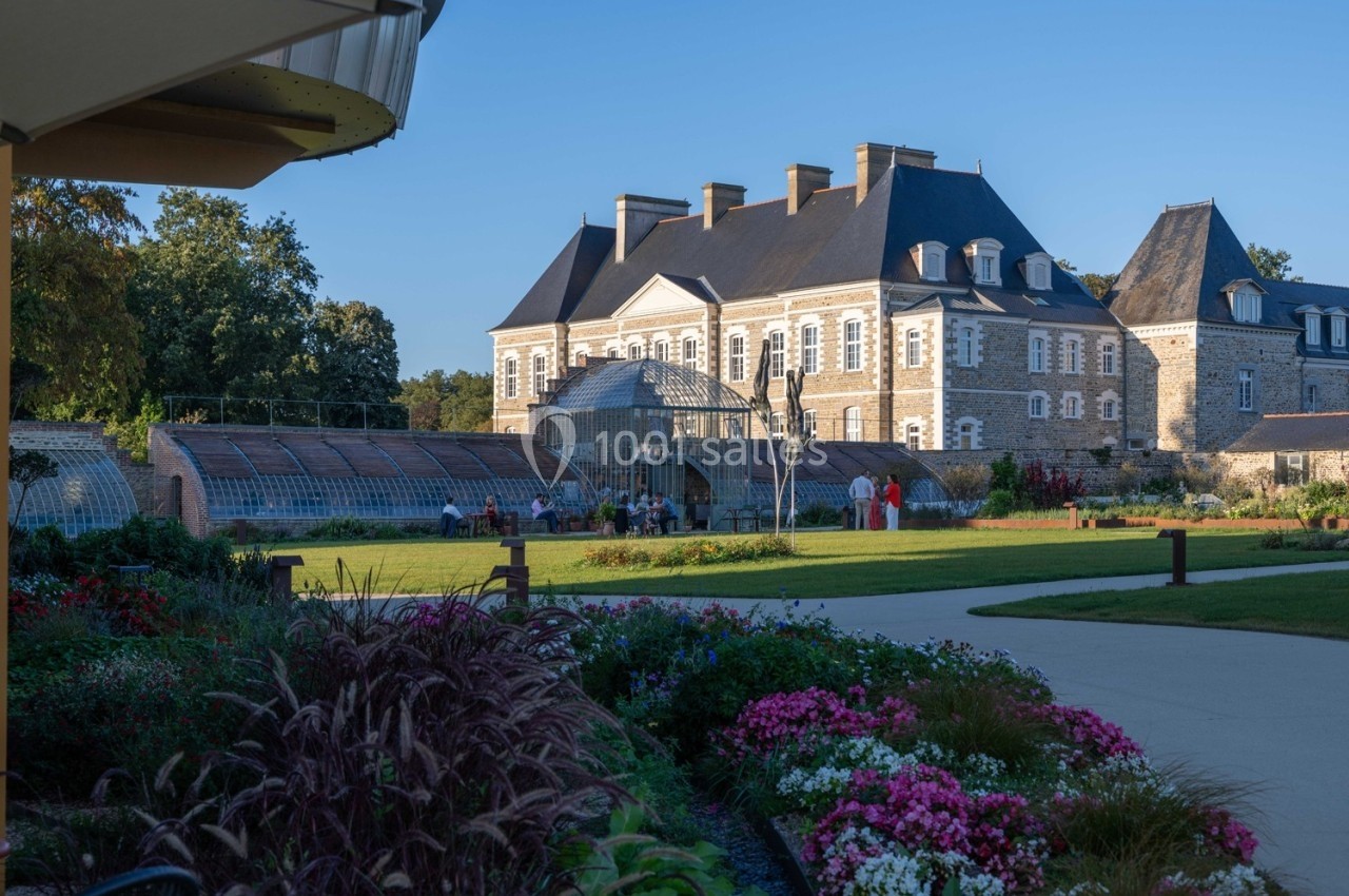 Jardin fleuri avec allée menant à un château en pierre et une serre, sous un ciel dégagé en fin de journée.