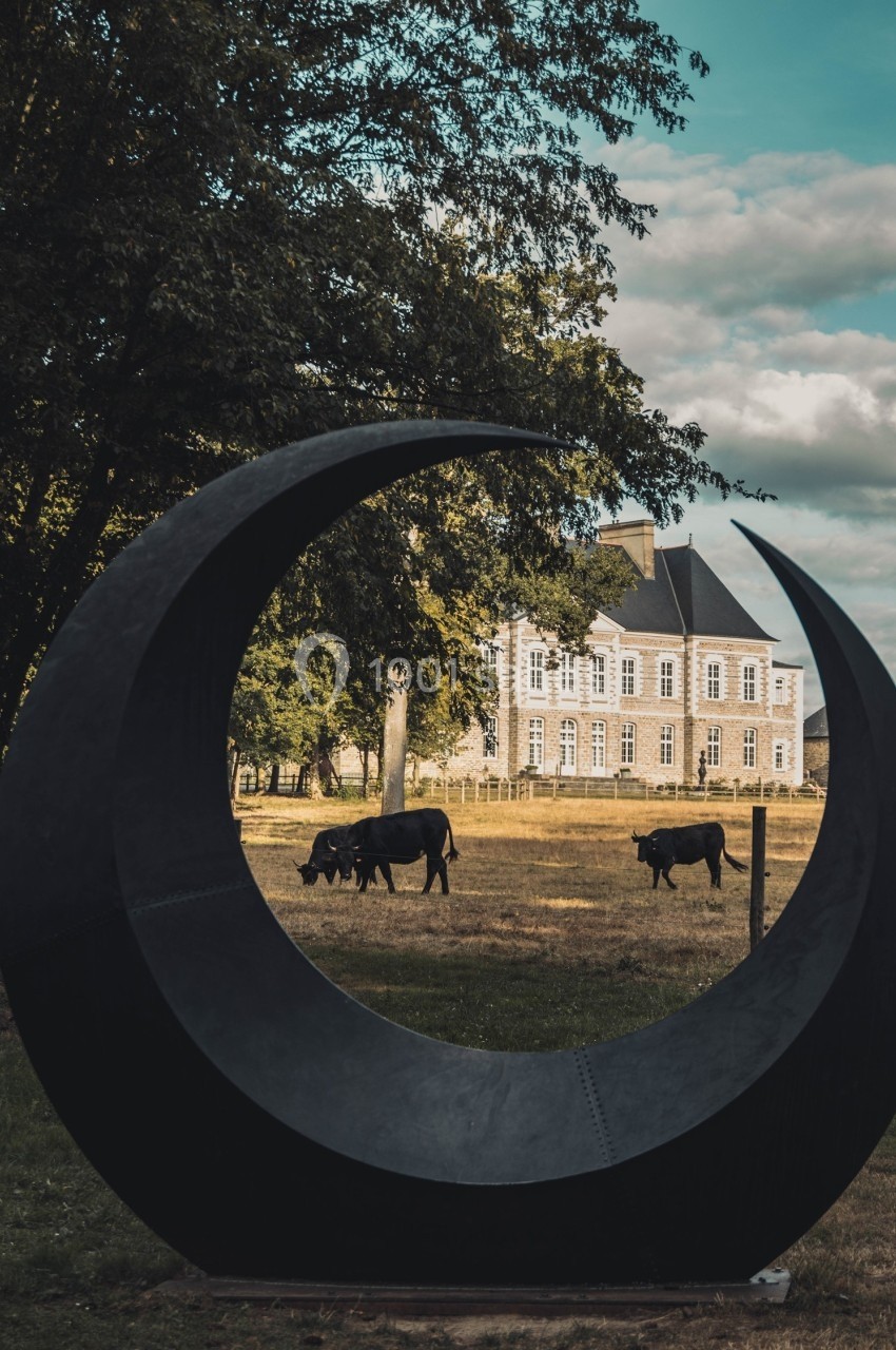 Sculpture en forme de croissant noir encadrant des vaches paissant devant un manoir entouré d'arbres.