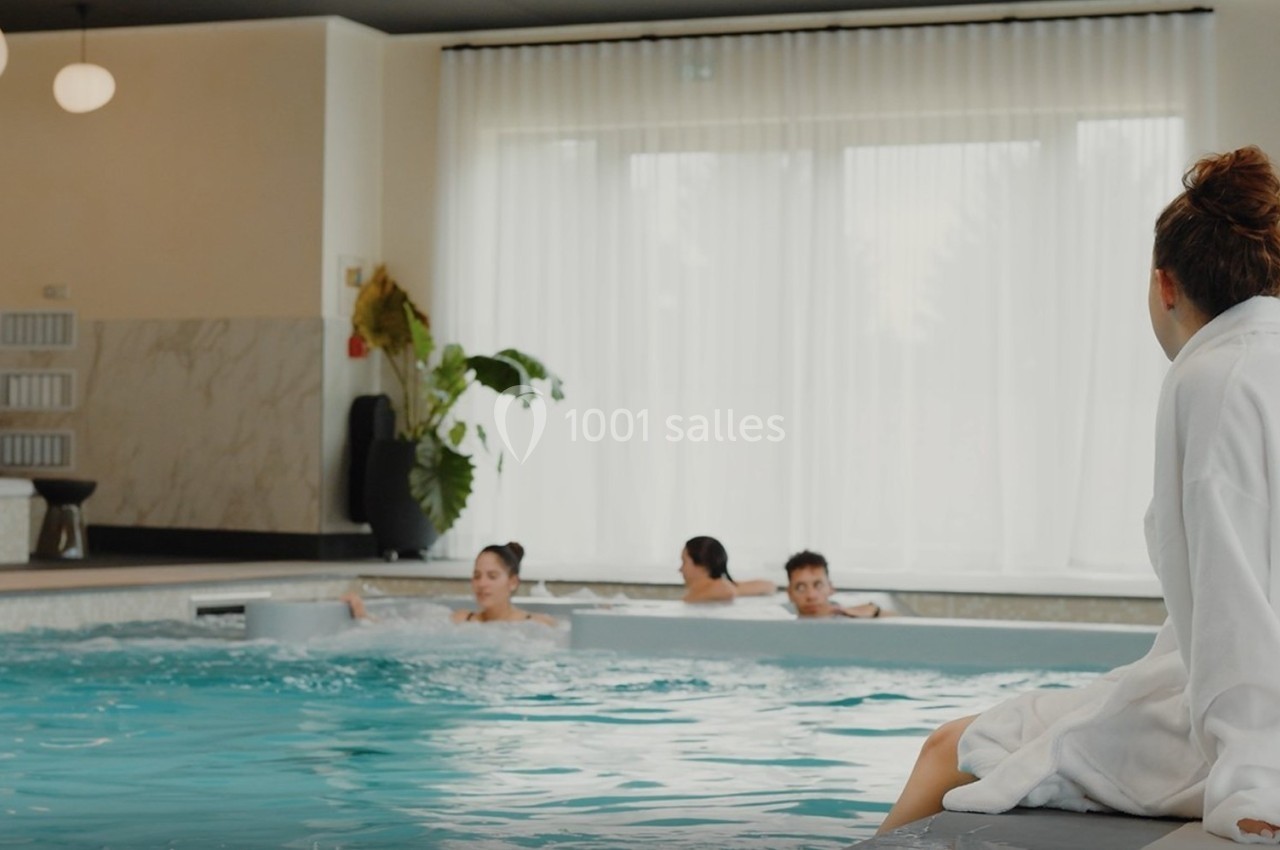 Une femme en peignoir assise au bord d'une piscine intérieure observe trois personnes nageant.