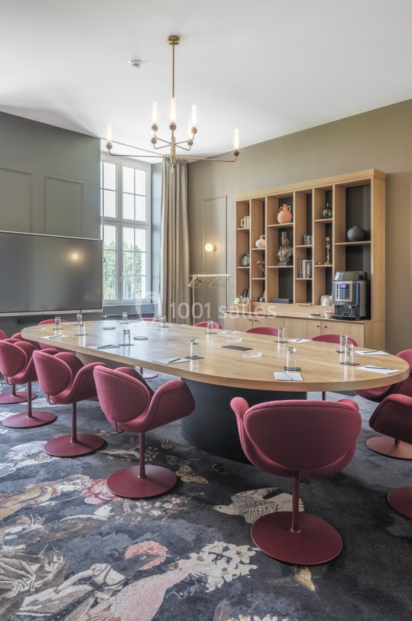 Salle de réunion lumineuse avec une grande table ovale en bois, chaises rouges, étagères et tableau blanc.