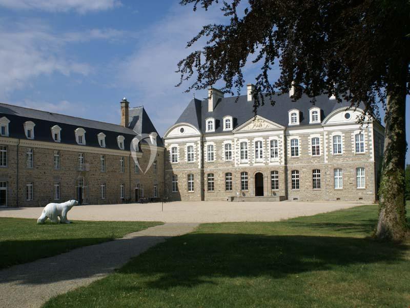 Façade d'un château en pierre avec toit en ardoise, entouré d'une cour gravillonnée et d'un espace vert.