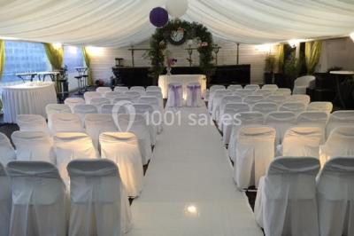 Décor champêtre avec fauteuils, tapis, grande horloge et lanternes sous un arbre, ambiance vintage en extérieur.