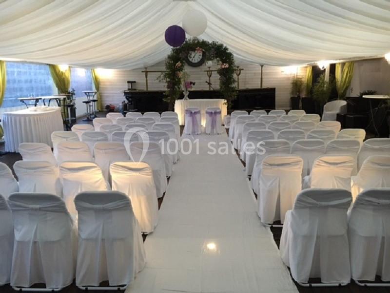 Salle de cérémonie décorée avec des chaises blanches, une allée centrale et une arche ornée de fleurs et de verdure.