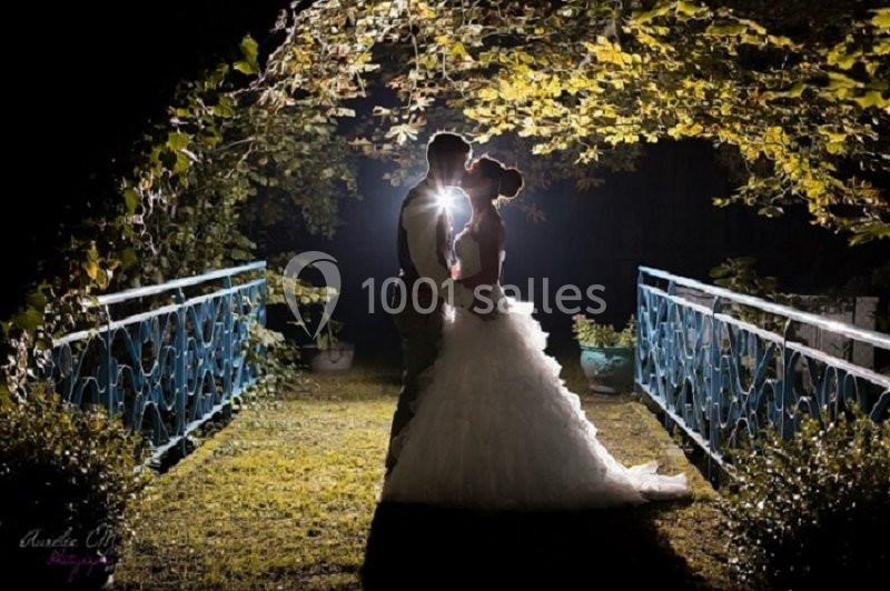 Un couple en tenue de mariage s'embrasse sur un pont éclairé, entouré de feuillage dans une ambiance nocturne.
