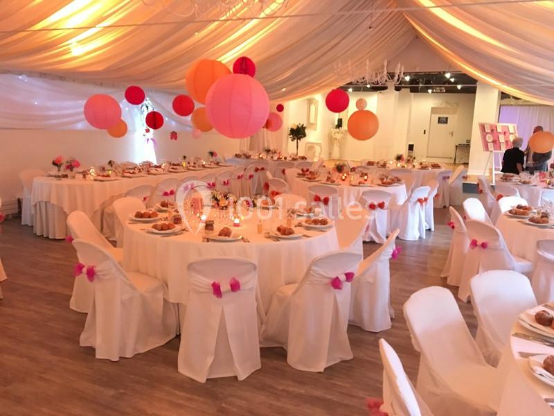 Salle de réception décorée avec des nappes blanches, des chaises ornées de rubans roses et des lanternes suspendues.