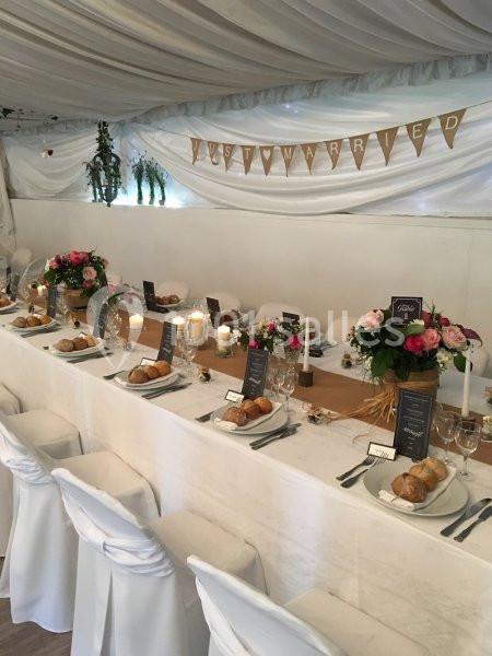 Table de réception décorée avec des fleurs, bougies et vaisselle, sous une tente avec une bannière ’Just Married’.