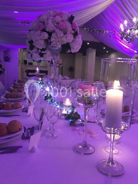 Table de réception élégamment décorée avec des fleurs roses, bougies allumées et vaisselle raffinée sous un éclairage violet.