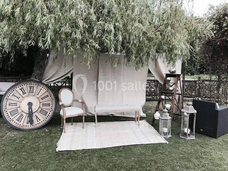 Décor champêtre avec fauteuils, tapis, grande horloge et lanternes sous un arbre, ambiance vintage en extérieur.