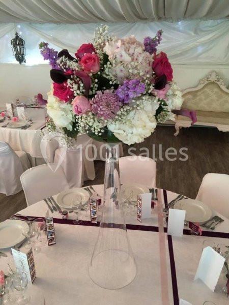 Centre de table floral avec roses, pivoines et gypsophiles dans un vase en verre haut, décorant une salle de réception.