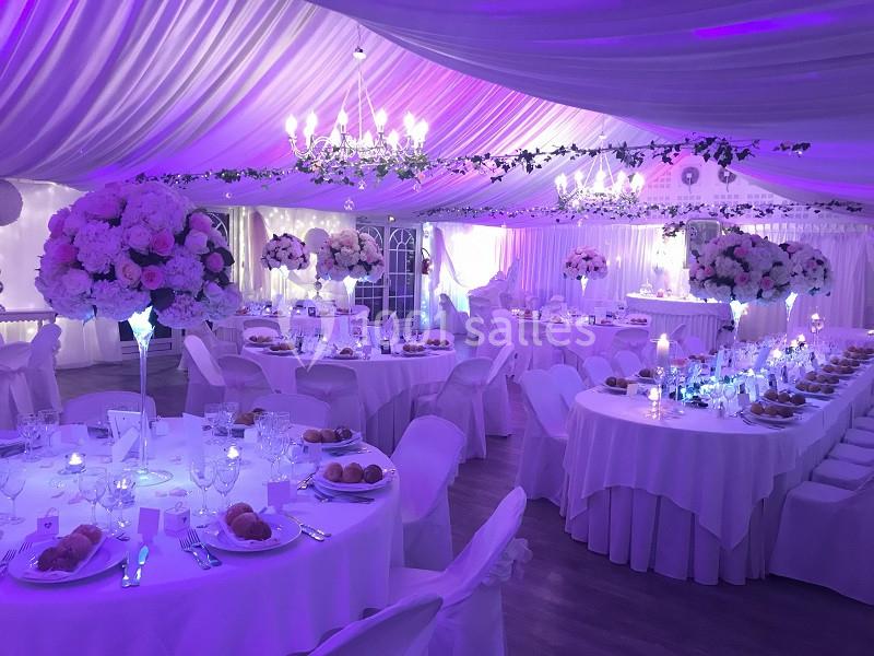 Salle de réception décorée avec des tables rondes, nappes blanches, chandeliers et éclairage violet.