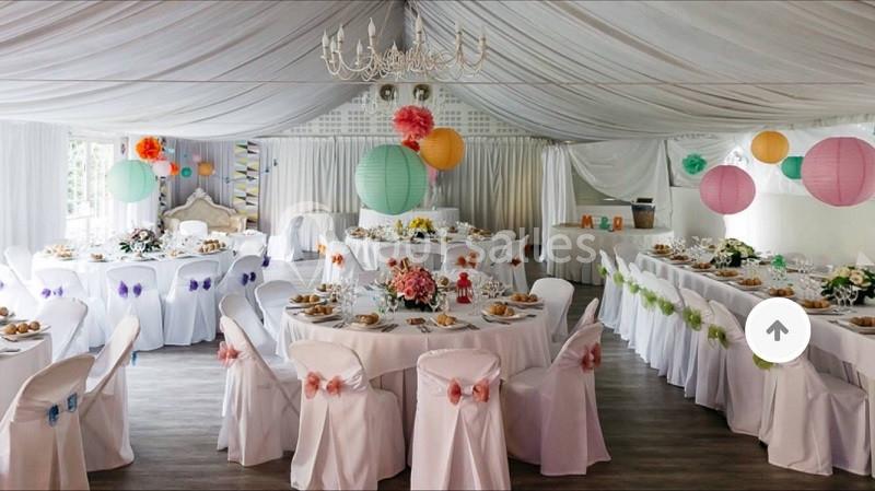 Salle de réception décorée avec des tables rondes, nappes blanches, fleurs colorées et lanternes suspendues.