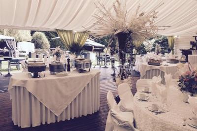 Décor champêtre avec fauteuils, tapis, grande horloge et lanternes sous un arbre, ambiance vintage en extérieur.