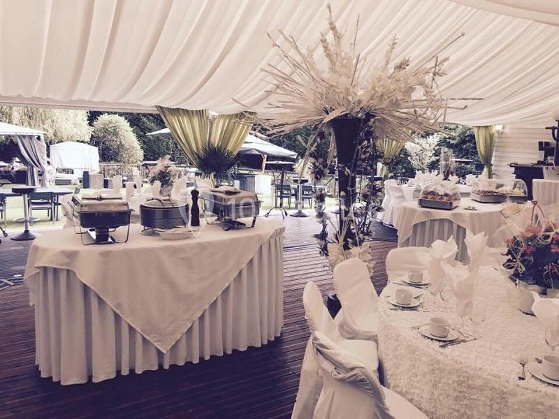 Salle de réception sous une tente avec tables dressées, nappes blanches et décorations florales, donnant sur un jardin.