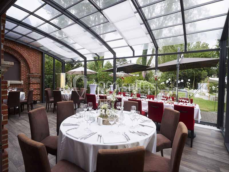 Salle de réception lumineuse avec tables dressées, chaises en tissu, baies vitrées et vue sur un jardin verdoyant.