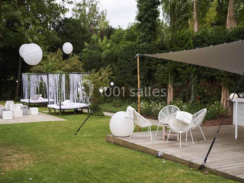 Espace extérieur aménagé avec chaises blanches, lits de repos, lanternes suspendues et végétation en arrière-plan.