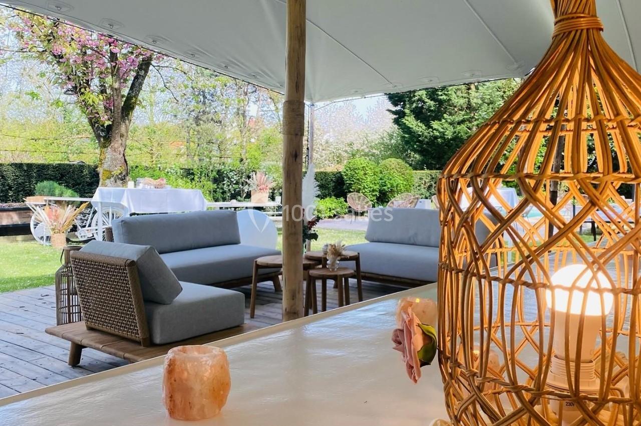 Espace extérieur aménagé avec des canapés, une table en bois et une lanterne en rotin sous une tente blanche.