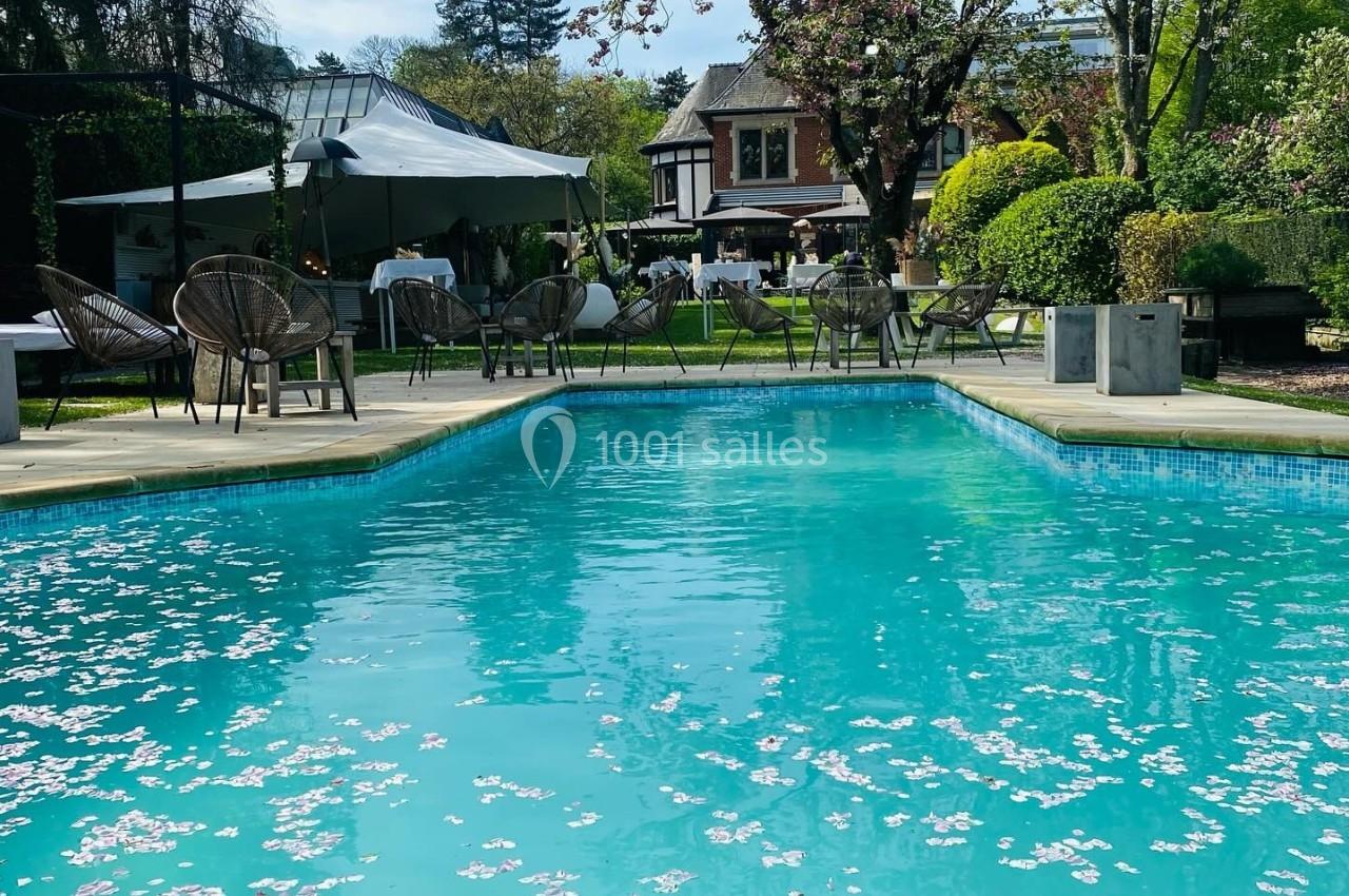 Piscine extérieure avec pétales de fleurs flottant à la surface, entourée d'arbres et d'une maison en arrière-plan.