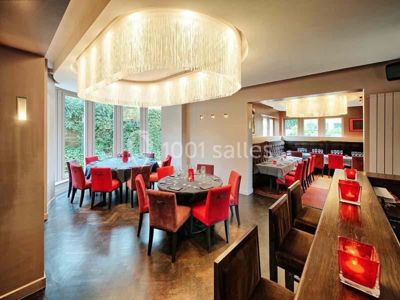 Salle de restaurant élégante avec tables dressées, chaises rouges, grandes baies vitrées et lustres modernes.