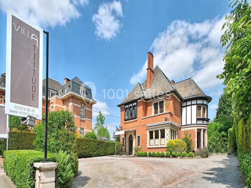 Façade d'une grande maison en briques avec toit en ardoise, entourée de verdure, et un panneau indiquant ’Villa Attitude’.