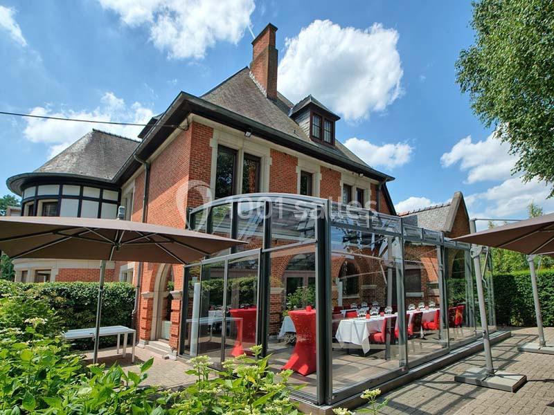 Maison en briques rouges avec une véranda vitrée aménagée pour un repas, entourée de verdure et de parasols.