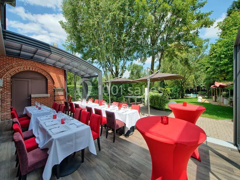 Terrasse aménagée avec tables dressées, chaises rouges, parasols et vue sur un jardin arboré par temps ensoleillé.