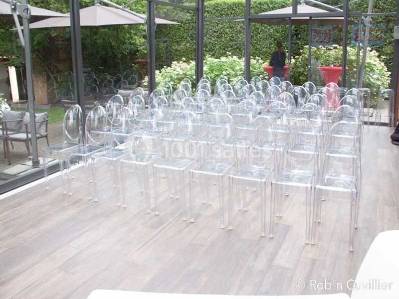 Rangées de chaises transparentes alignées dans une salle lumineuse avec baies vitrées donnant sur un jardin.