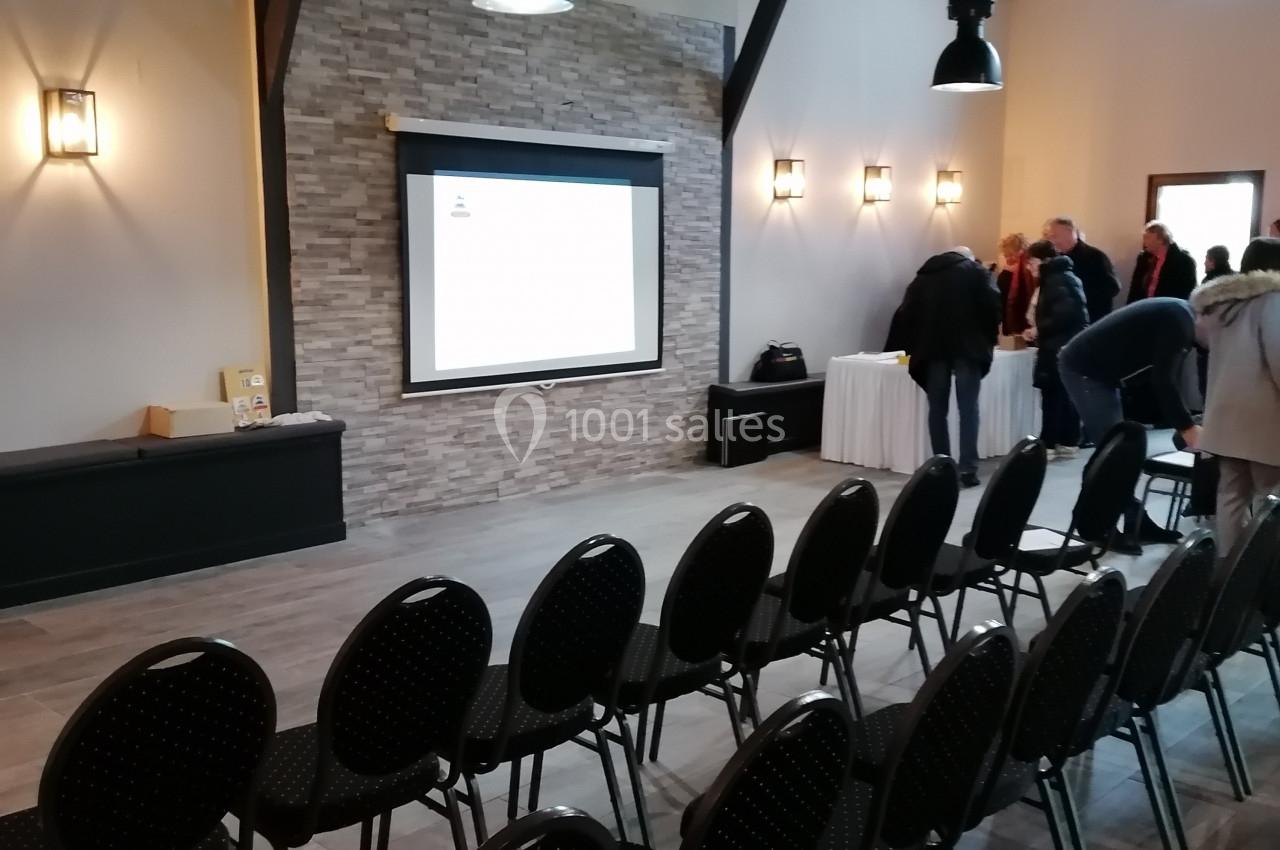 Salle de conférence avec des rangées de chaises noires, un écran de projection allumé et quelques personnes debout.