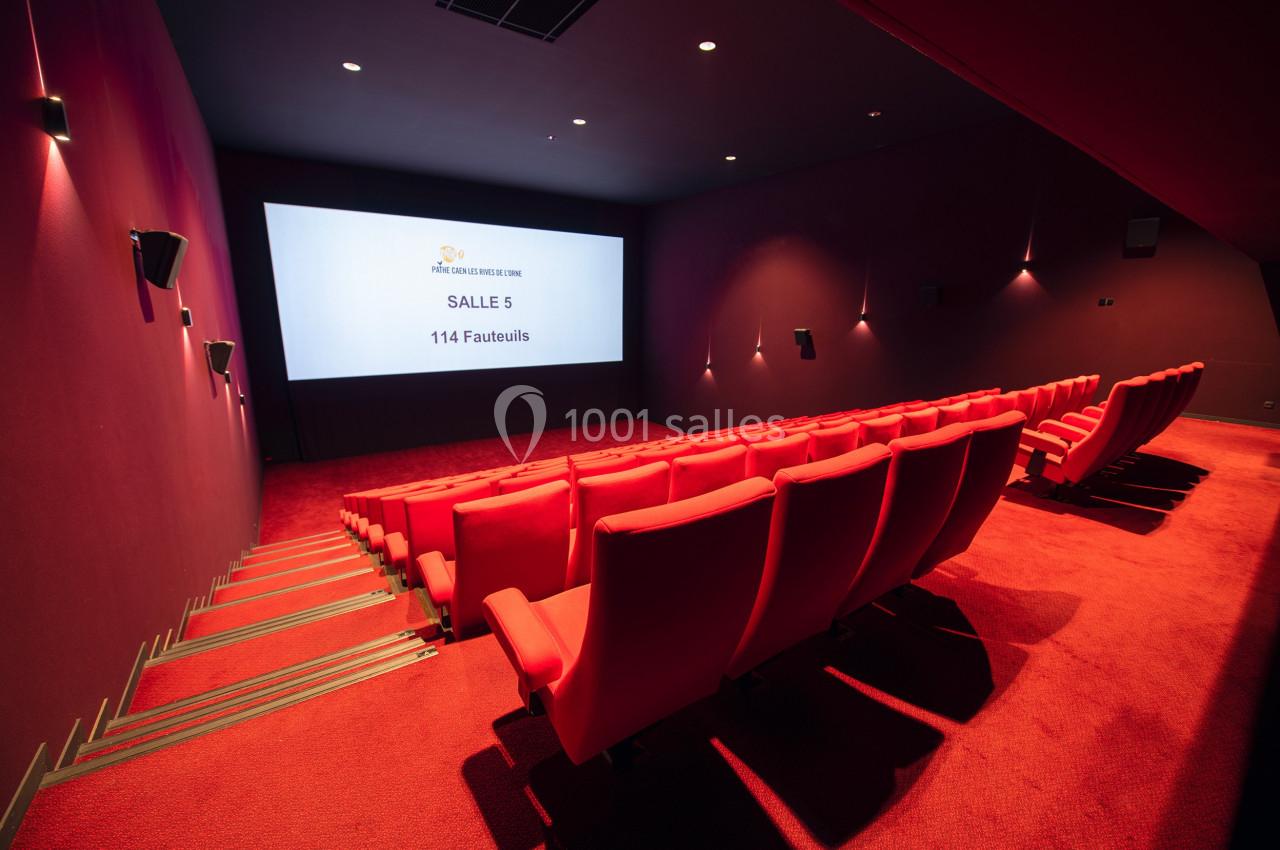 Salle de cinéma vide avec des sièges rouges, écran allumé affichant ’Salle 5, 114 fauteuils’, éclairage tamisé.