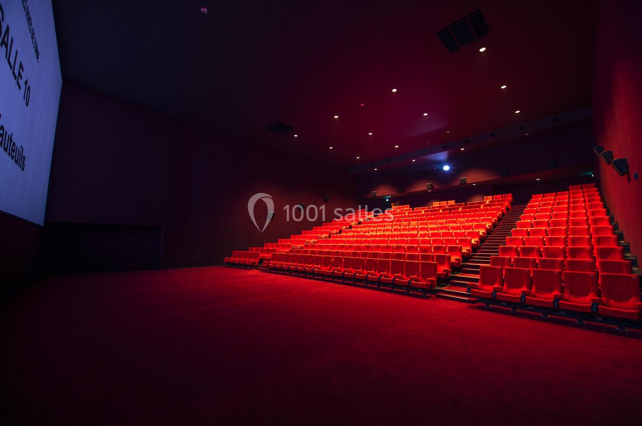 Salle de cinéma vide avec rangées de sièges rouges et écran blanc sur la gauche.