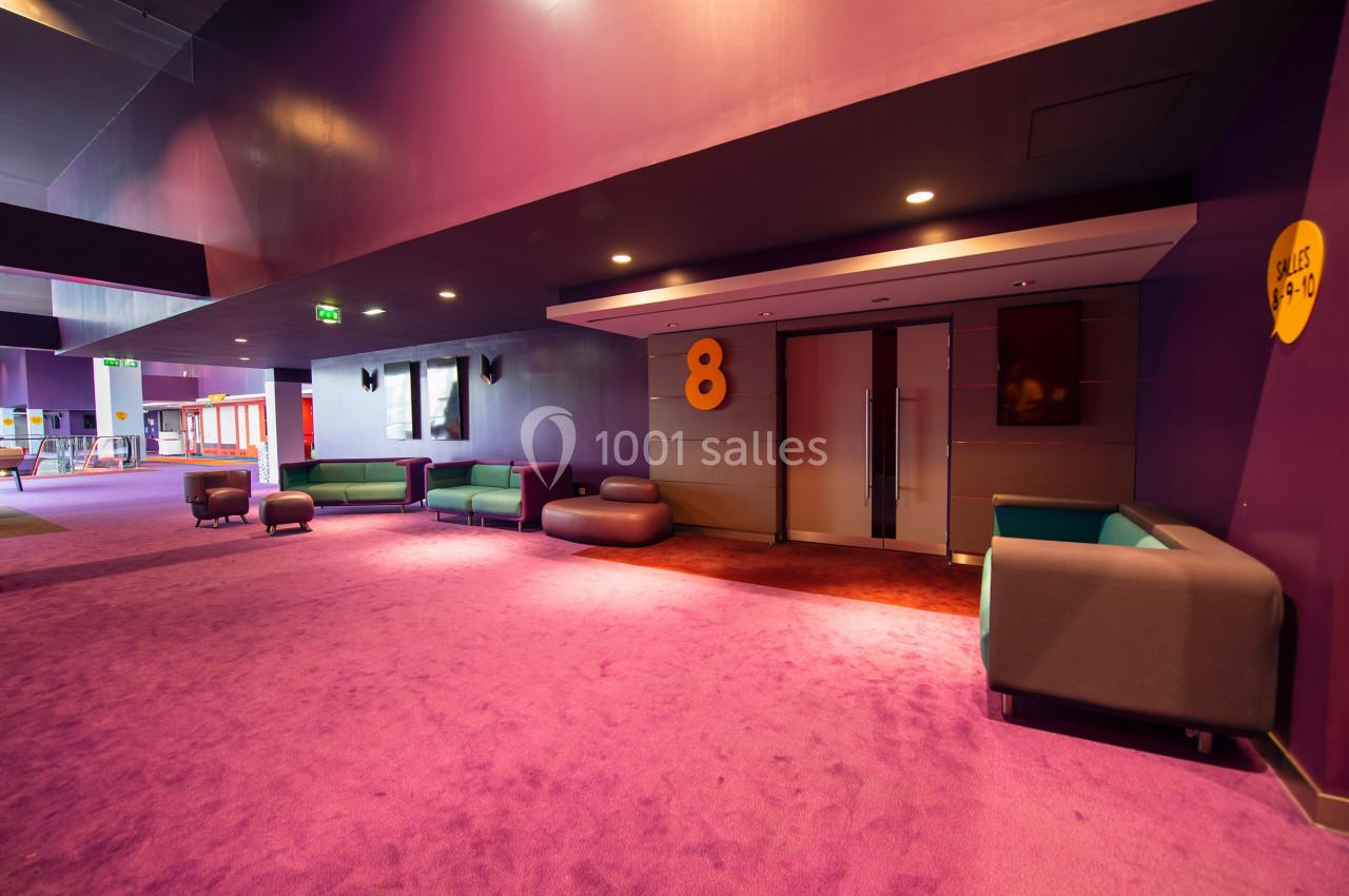 Couloir d'un cinéma avec moquette violette, canapés modernes, et porte numérotée ’8’ menant à une salle.