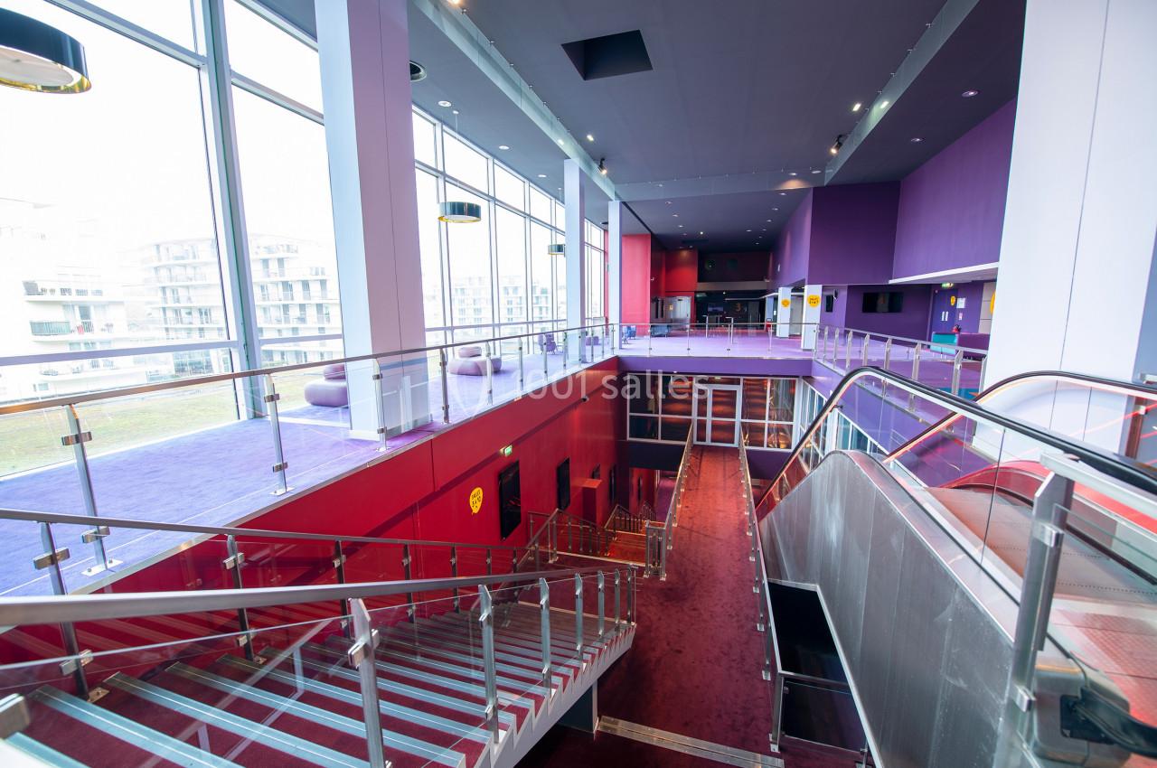 Intérieur d'un bâtiment moderne avec escalier, rambardes en métal, murs colorés et grandes baies vitrées.
