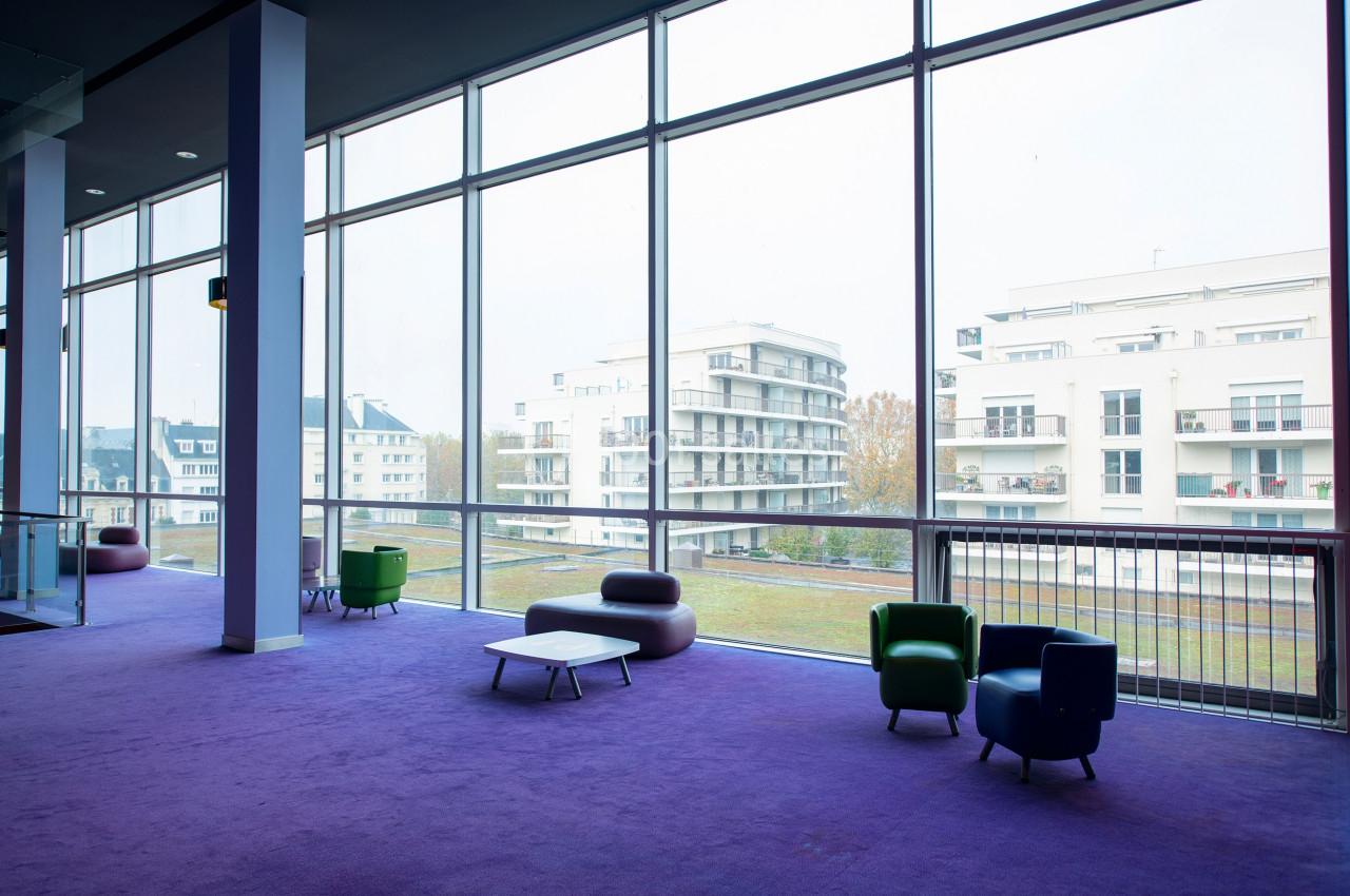 Espace intérieur lumineux avec moquette violette, fauteuils colorés et grandes baies vitrées donnant sur des immeubles…