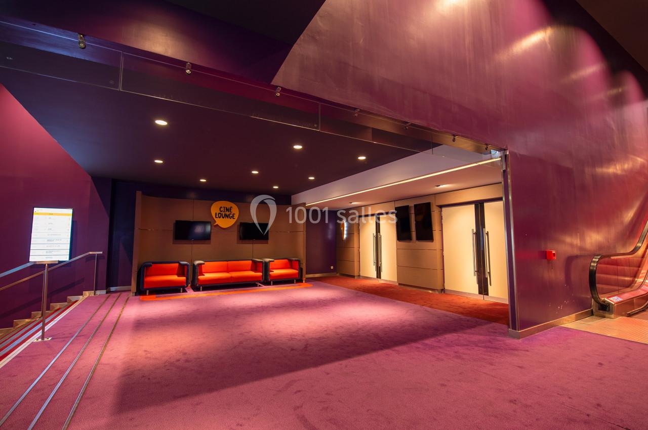 Hall d'entrée d'un cinéma avec moquette violette, sièges rouges, écrans muraux et escalier roulant à droite.