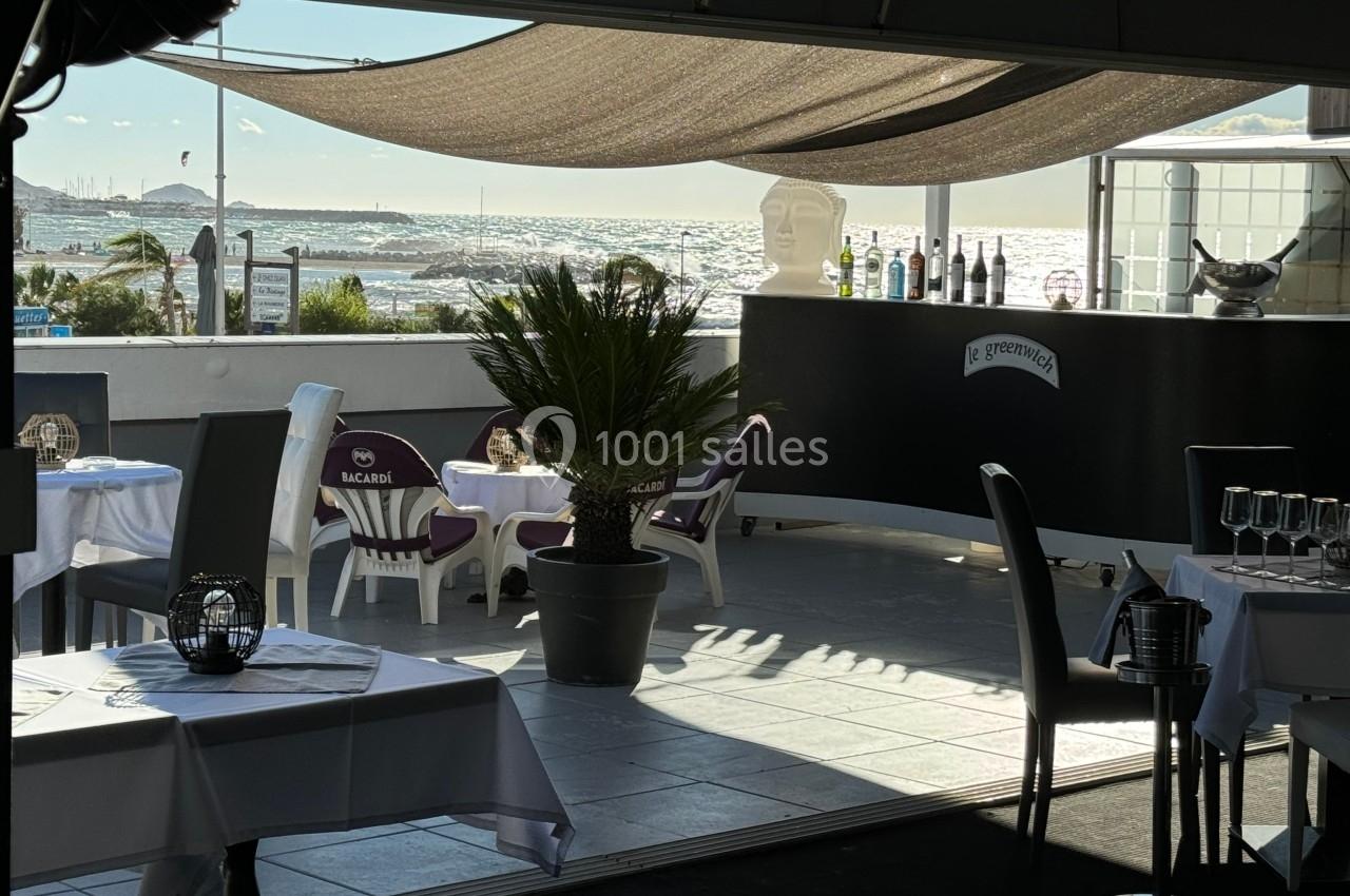 Terrasse d'un restaurant avec vue sur la mer, tables dressées, palmier en pot et comptoir avec bouteilles exposées.