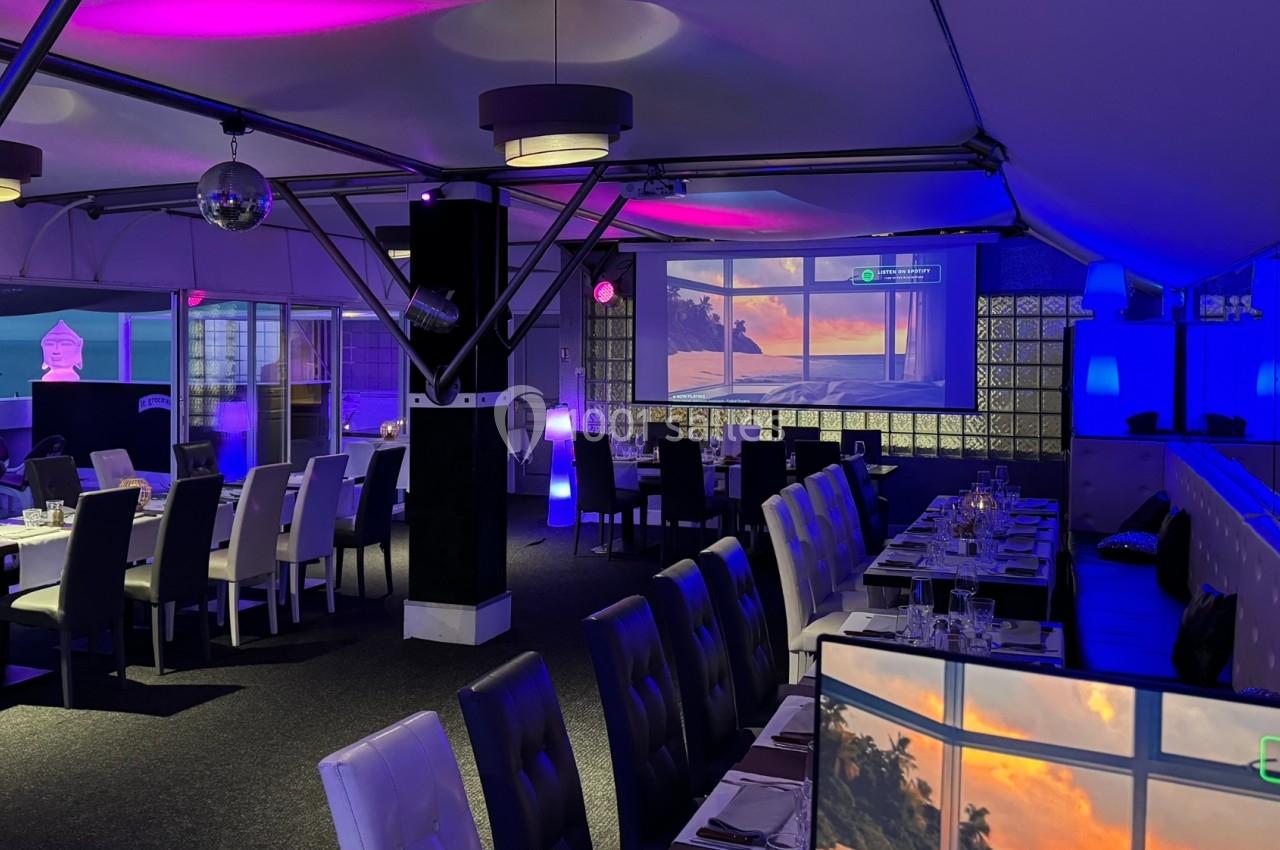 Salle de restaurant moderne avec éclairage violet, tables dressées et vue sur un coucher de soleil à travers une baie vitrée.