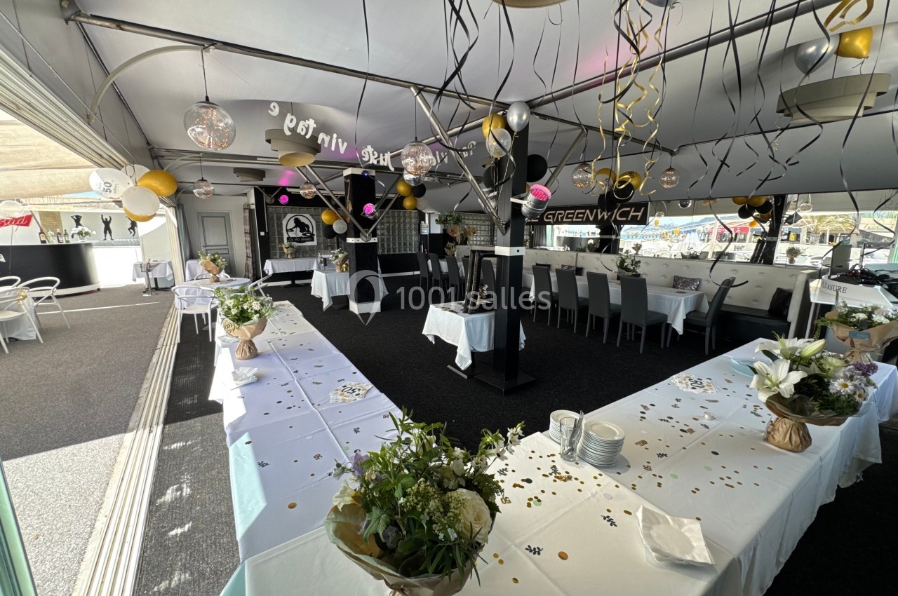 Salle décorée pour une fête, avec tables dressées, ballons, guirlandes et fleurs dans un espace lumineux.